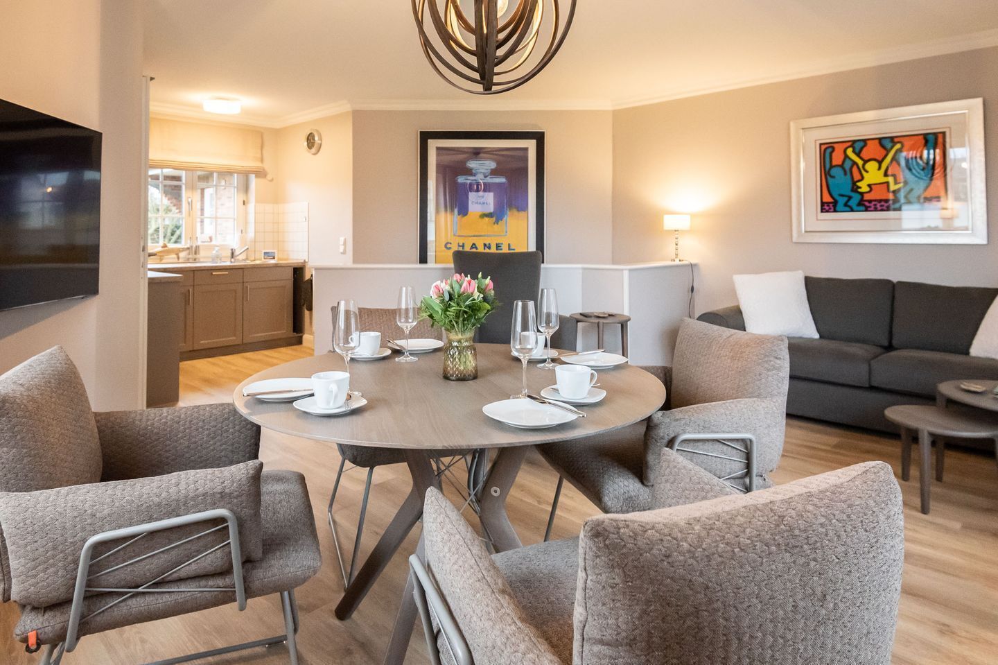 Una sala moderna con una mesa de comedor redonda preparada para cuatro personas, sillas tapizadas en gris, un sofá gris y un televisor de pantalla plana montado en la pared.