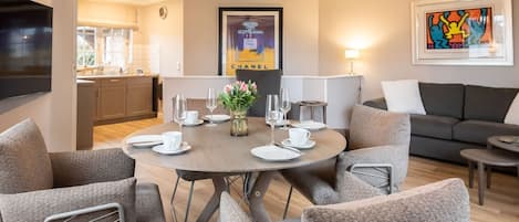 Ein modernes Wohnzimmer mit einem runden Esstisch für vier Personen, grauen Polsterstühlen, einem grauen Sofa und einem flachbildfernseher an der Wand.