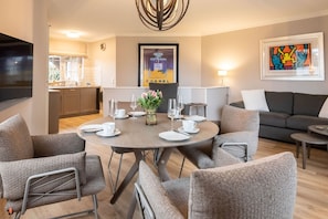 A modern living room with a round dining table set for four, grey upholstered chairs, a grey sofa, and a flat-screen TV mounted on the wall. - Achter Dünem, Whg. 4 - Strandnahe 3-zimmer Ferienwohnung auf 70 m² Wohnfläche, für bis zu 4 Personen (Wenningstedt-Braderup (Sylt))