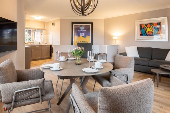 A modern living room with a round dining table set for four, grey upholstered chairs, a grey sofa, and a flat-screen TV mounted on the wall.