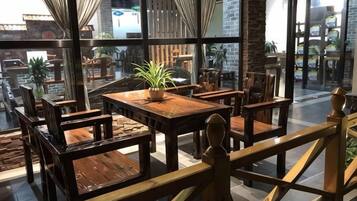 A restaurant with wooden tables and chairs, a potted plant, and large windows offering a view of the street.