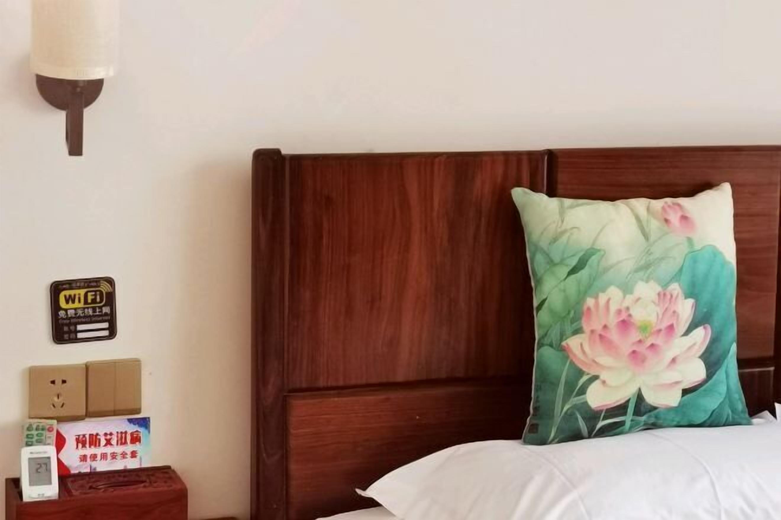 Ett hotellrum med en sänggavel i trä, en prydnadskudde med blommönster, ett nattduksbord med en klocka, en skylt som visar att det finns Wi-Fi och en skylt med kinesiska tecken.