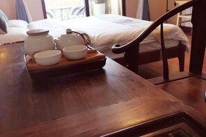 Una mesa de madera con una bandeja que contiene teteras y tazas, una cama con sábanas blancas y una ventana con cortinas al fondo.