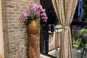 Un muro de ladrillo, un florero de madera con flores y una ventana con cortinas.