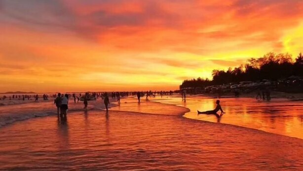 A sunset over a beach with people walking and sitting on the sand.