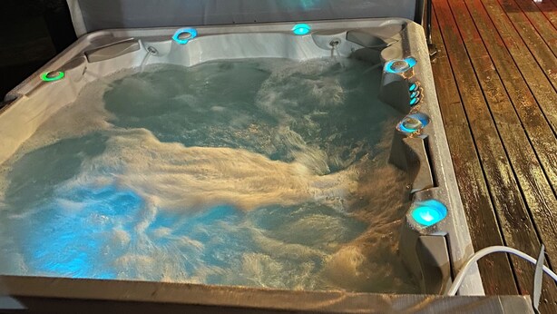 Ein Whirlpool mit mehreren Düsen, umgeben von einer Holzterrasse und einem überdachten Sitzbereich mit Tischen und Stühlen.