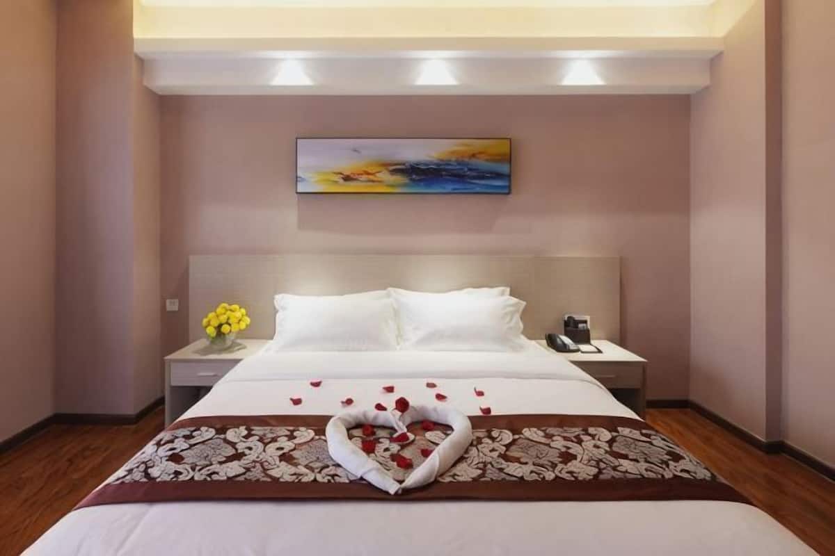 Quarto de hotel com uma cama grande, uma mesa de cabeceira com luminária, um quadro na parede e um arranjo de flores sobre a cama.