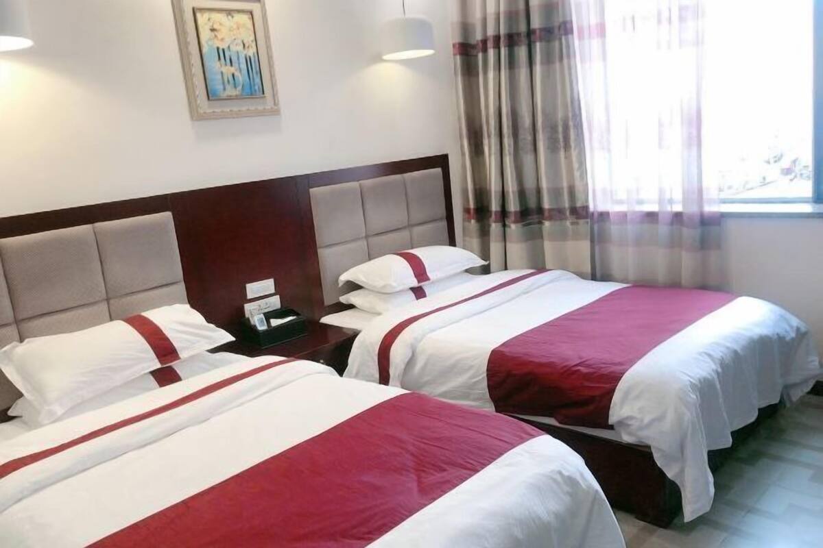 Quarto de hotel com duas camas, cada uma com roupa de cama branca e vermelha, uma cabeceira de madeira, uma mesa de cabeceira e um quadro na parede.