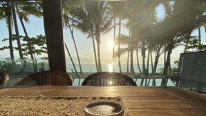 Ett träbord med en vävd bordsduk och en vit kopp, med utsikt över en strand med palmer och havet, under en klar himmel.