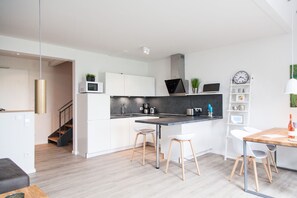 Une cuisine moderne avec des armoires blanches, un comptoir noir et une cuisinière intégrée. On y trouve deux tabourets et une table en bois sur laquelle est posée une bouteille.