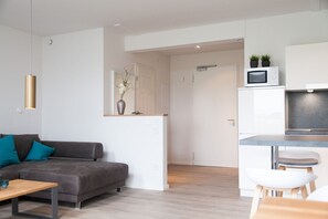 Ein modernes Wohnzimmer mit grauem Sofa, türkisfarbenen Kissen, einem Holztisch und einer weißen Kommode mit Mikrowelle.