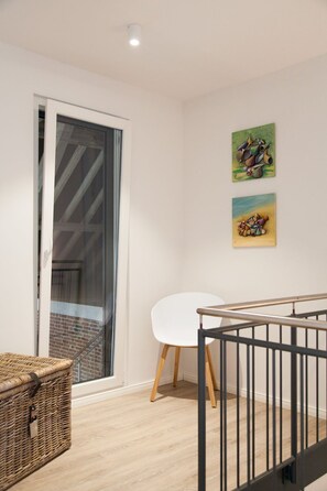 A room with a sliding glass door leading to a balcony, a white table with a chair, and two artworks on the wall.