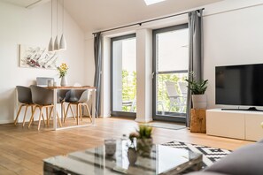 A modern living room with a dining area, large sliding glass doors to a balcony, a flat-screen TV, and a minimalist decor style.