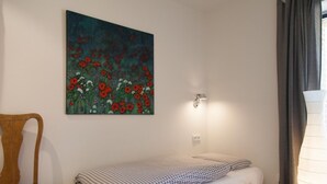Une chambre avec un lit simple, une chaise en bois, une lampe fixée au mur et un tableau représentant des fleurs.