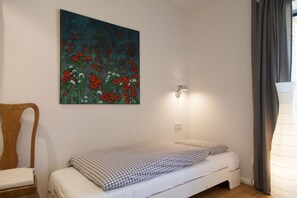 Ein Schlafzimmer mit einem Einzelbett, einem hölzernen Stuhl, einer Wandleuchte und einem Blumengemälde.