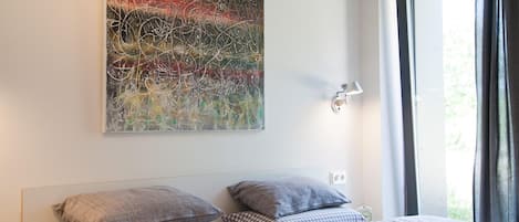 A bedroom with two beds, a large abstract painting on the wall, a bedside lamp, and a window with curtains.