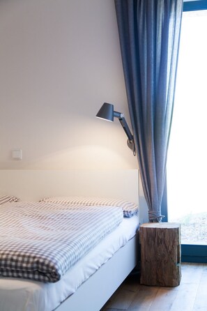 Un dormitorio con una cama, una lámpara de noche, un taburete de madera y una ventana con cortinas.