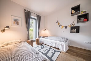 Un dormitorio con dos camas, una ventana con cortinas, una lámpara de pared y un pequeño estante con libros y un juguete.