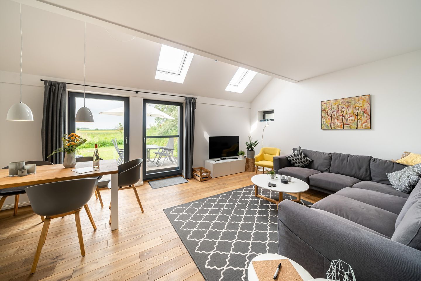 A modern living room with a grey sofa, a dining table with chairs, a TV, and a large window with curtains.