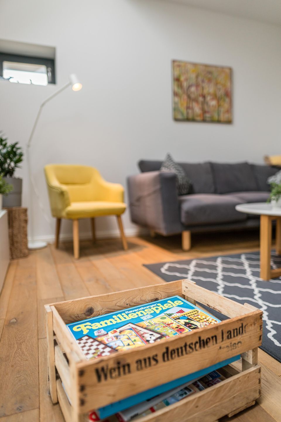 Une caisse en bois contenant des livres, sur laquelle on peut lire l’inscription « Wein aus deutschen Land », se trouve dans un salon avec un fauteuil jaune, un canapé gris et un tapis noir et blanc.