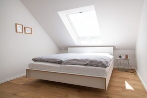 Ein Schlafzimmer mit schrägem Dach, einem Oberlicht, einem Bett mit Karomuster-Bettwäsche, einem Nachttisch mit Radio und einem kleinen Stuhl mit Drahtgestell.