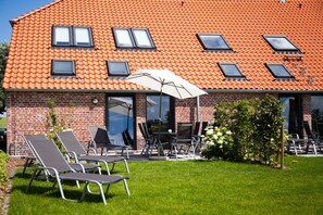 Ein Ziegelhaus mit Ziegeldach, eine Terrasse mit Stühlen und Tisch sowie ein Garten mit einem kleinen Baum und Blumen.