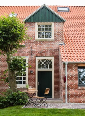 Ein Backsteinhaus mit rotem Ziegeldach, einem zentralen Eingang mit Glastür und einer kleinen Außenbestuhlung mit Tisch und zwei Stühlen.