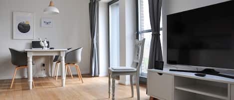 A modern living room with a large TV, a dining area with a white table and chairs, and large windows with curtains.