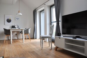Ett modernt vardagsrum med en stor TV, en matplats med ett vitt bord och stolar samt stora fönster med gardiner.