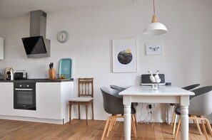 Eine moderne Küche mit weißen Schränken, einem schwarzen Ofen und einer Türkis-Arbeitsplatte. Es gibt einen Holzstuhl, einen Esstisch mit zwei Stühlen und eine Pendelleuchte.