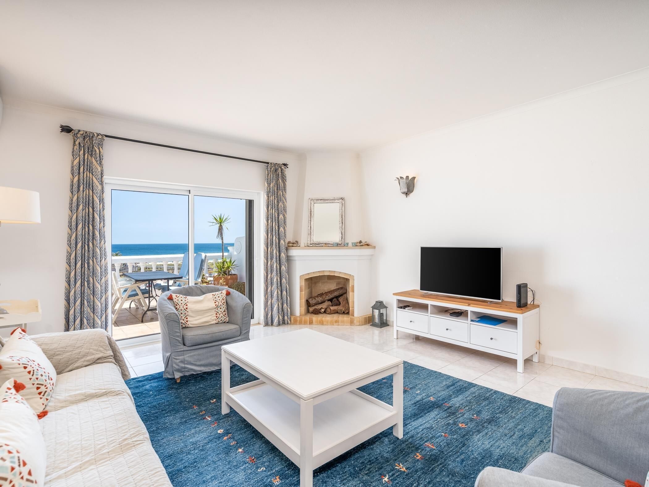 Un salon avec un téléviseur, des meubles blancs, une cheminée et une vue sur la mer.