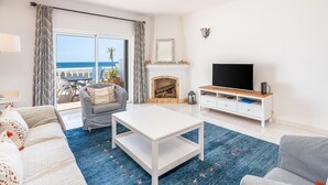 Sala de estar com televisão, móveis brancos, lareira e vista para o mar.
