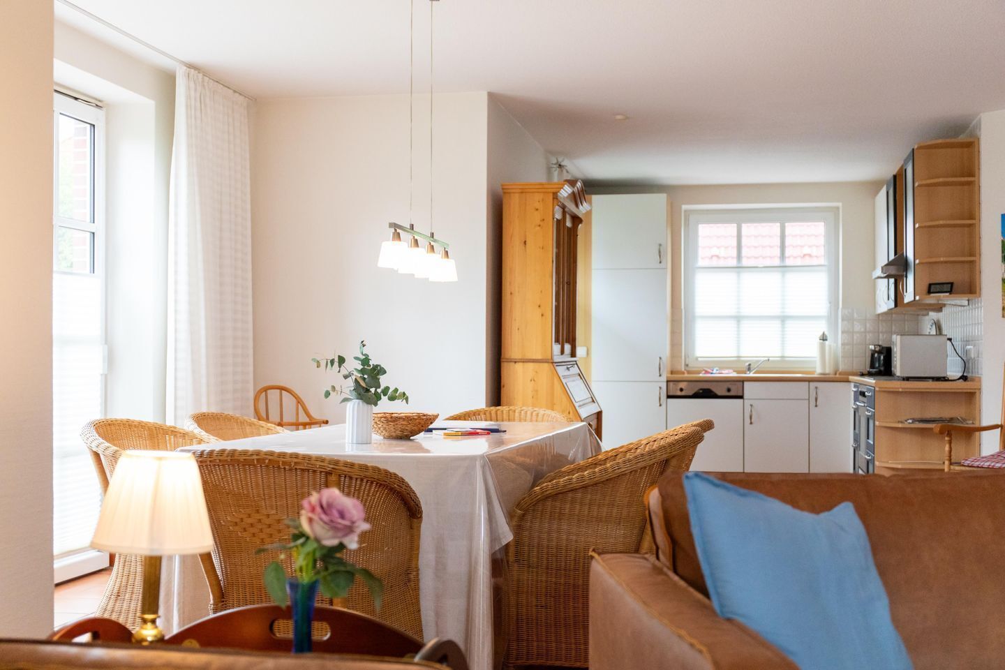 Un salon chaleureux avec un coin repas, des meubles en osier, une cuisine avec des placards en bois et une vue sur l’extérieur.