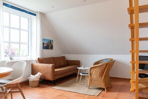 Ein gemütliches Wohnzimmer mit einer braunen Couch, einem Korbstuhl, einem kleinen runden Tisch und einem Fenster mit Blick auf Bäume.