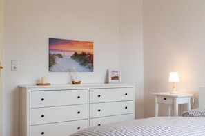 Ein Schlafzimmer mit Bett, einer Kommode mit Schubladen, einem Nachttisch mit Lampe und einem Wandbild mit Strandmotiv.