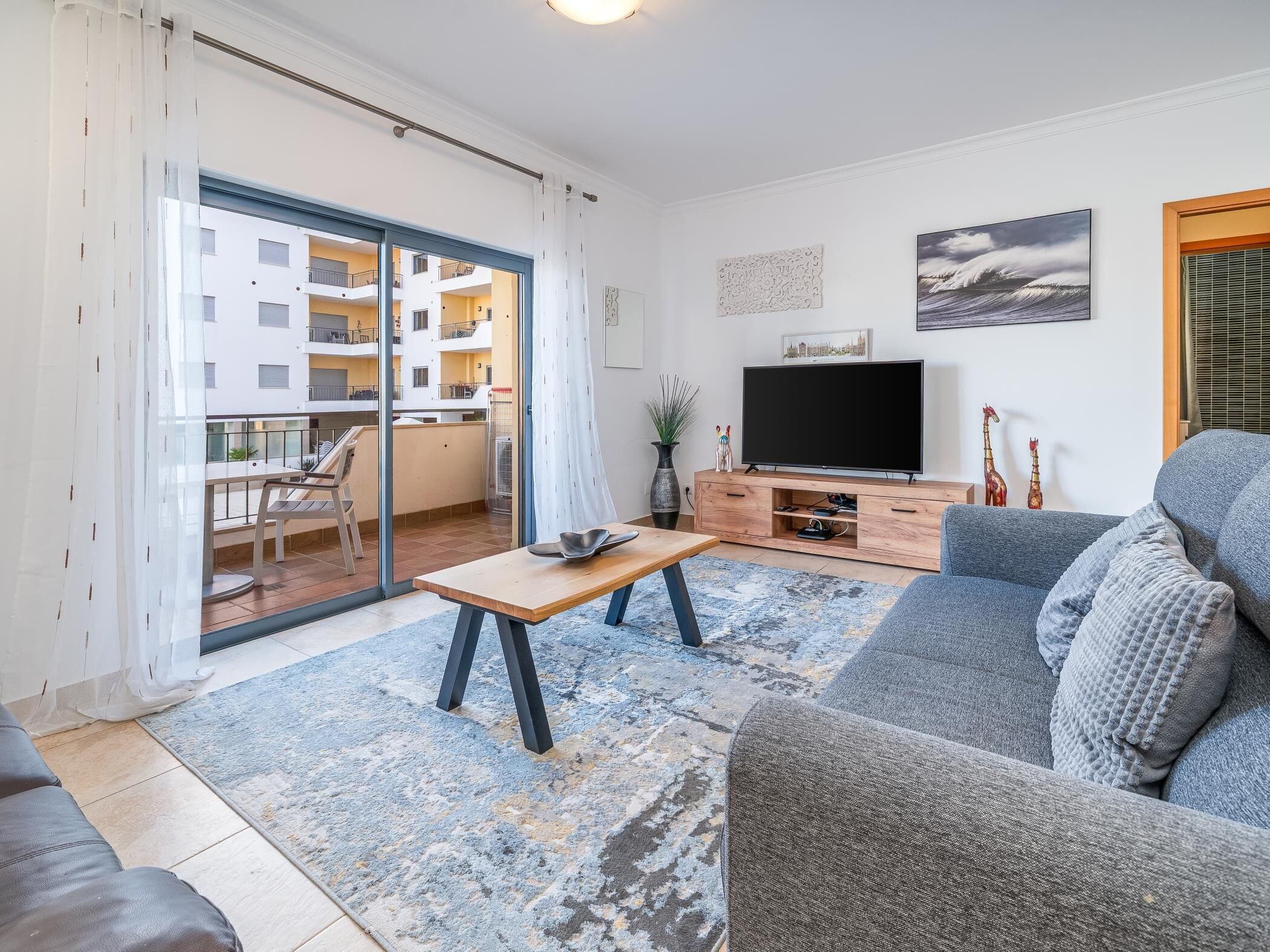 En moderne stue med en grå sofa, et træ sofabord og et fladskærms-tv på et træstel. Der er en balkon med udsigt til en hvid bygning og et spiseområde med et bord og stole.