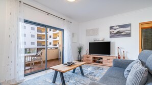 Ein modernes Wohnzimmer mit einem grauen Sofa, einem Holztisch und einem Flachbildfernseher auf einem Holzständer. Ein Balkon bietet Blick auf ein weißes Gebäude, und eine Essecke mit Tisch und Stühlen ist ebenfalls vorhanden.