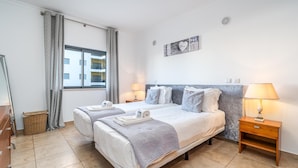 Ein Hotelzimmer mit zwei Betten, einem fensterbelaubten Fenster, einem Wandbild und einem Nachttisch mit Lampe.