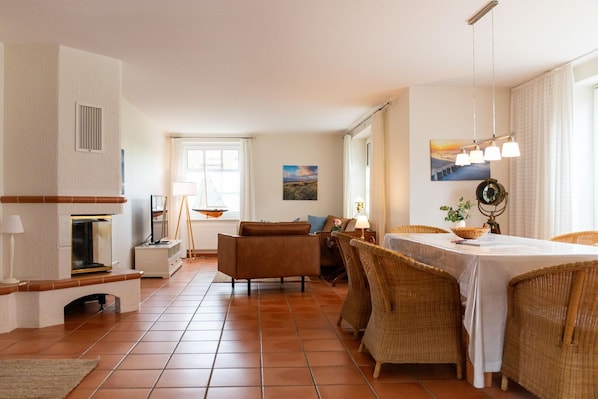 A spacious living room with a fireplace, a dining area with wicker furniture, and a view of the outdoors through large windows.