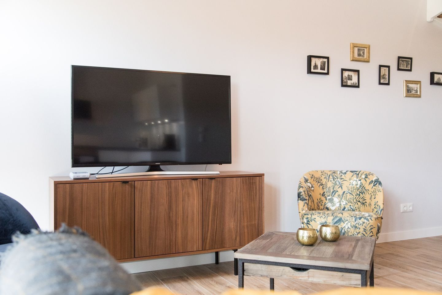 Uma sala de estar com televisão de tela plana sobre um armário de madeira, uma poltrona estampada e uma mesa de centro com objetos decorativos.