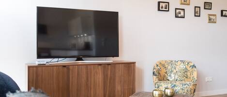 Uma sala de estar com televisão de tela plana sobre um armário de madeira, uma poltrona estampada e uma mesa de centro com objetos decorativos.