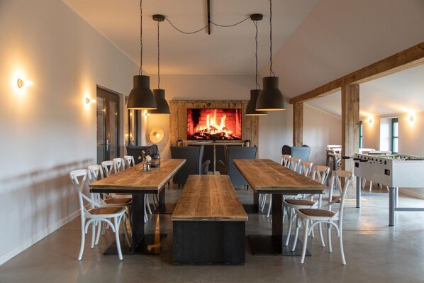 A spacious dining area with a long wooden table, white chairs, pendant lighting, and a fireplace.