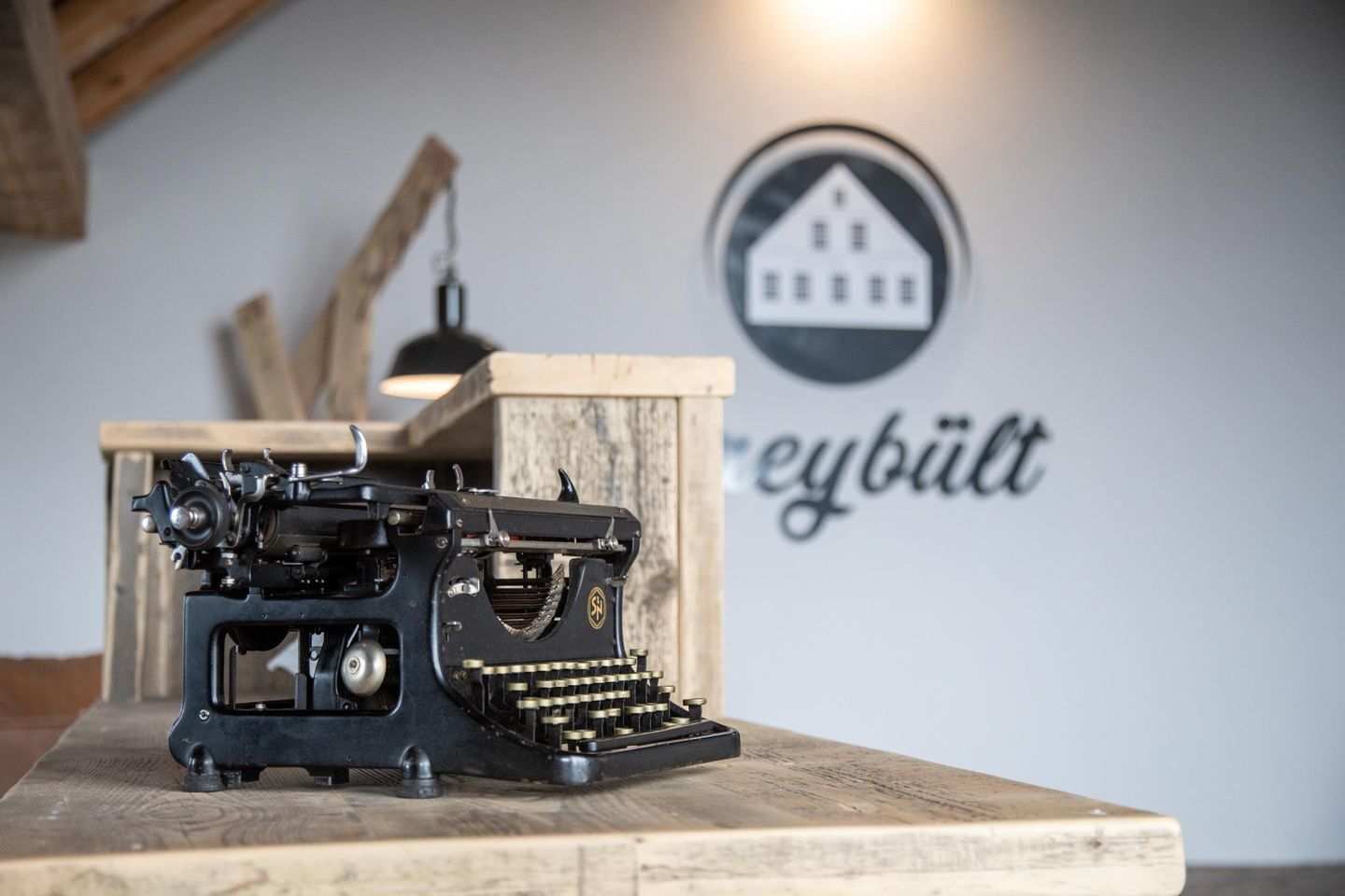 Une machine à écrire ancienne, posée sur une table en bois, avec un mur en arrière-plan sur lequel on peut lire le texte « reybütt ».