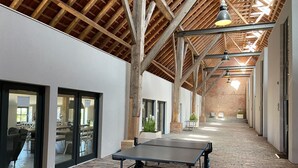 Ein Innenraum mit Tischtennisplatte, Holzbalken, Backsteinboden und großen Fenstern.