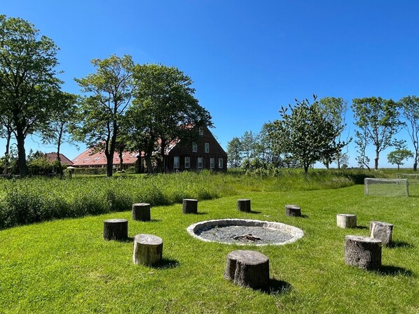 A grassy area with a circular stone feature, possibly a well or fire pit, surrounded by evenly spaced wooden posts. In the background, there is a large red-roofed building with multiple windows.