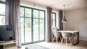 Ein modernes Zimmer mit Fernseher, Schreibtisch mit Stühlen, großem Fenster mit Vorhängen und einer Pendelleuchte.