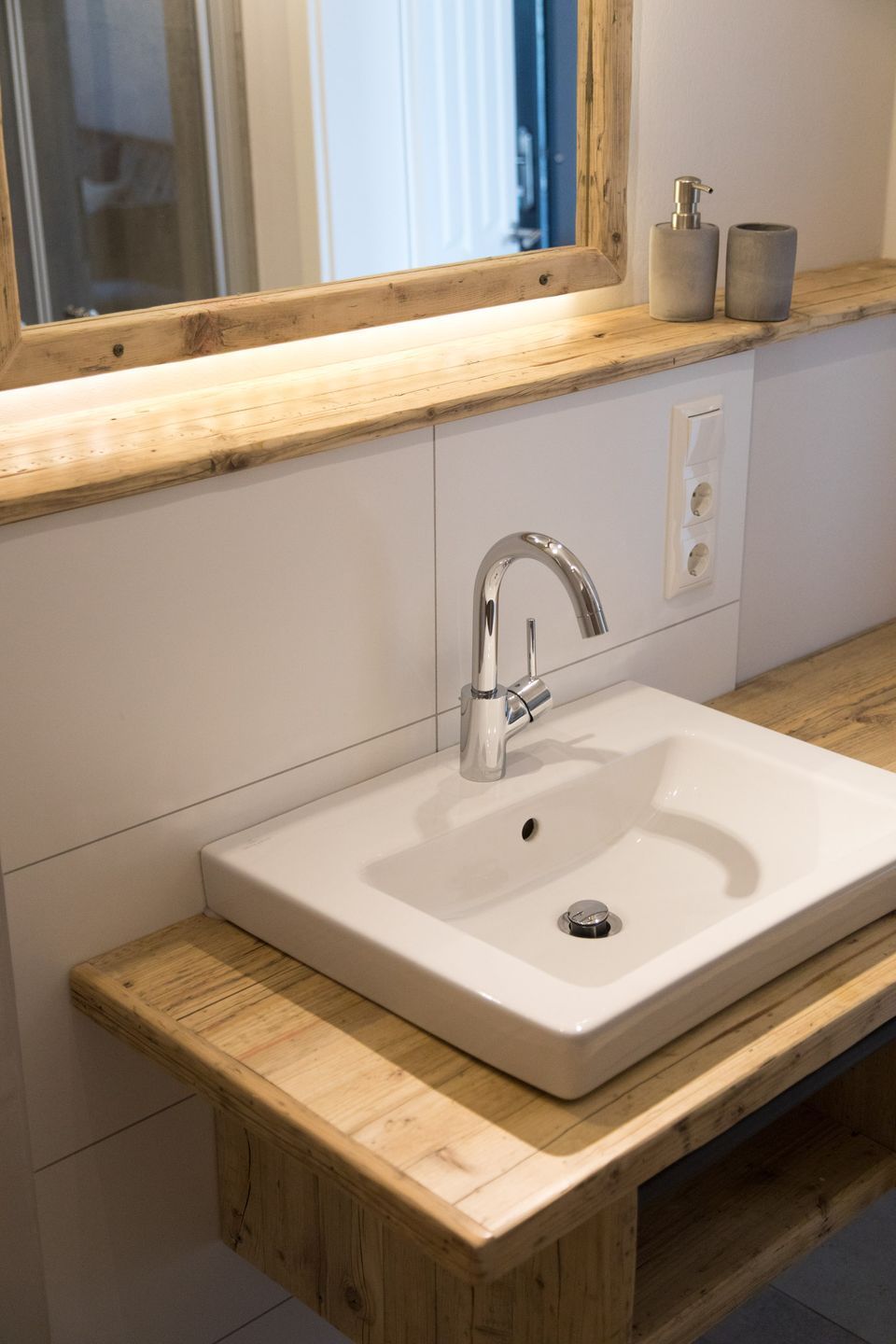 Un baño con tocador de madera, un lavamanos blanco, un grifo cromado y un espejo sobre el lavamanos.
