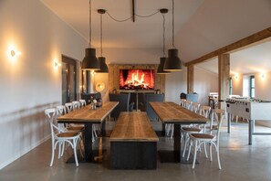 Un vaste espace dédié à la salle à manger, avec une longue table en bois, des chaises blanches, des luminaires suspendus et une cheminée.