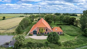 A large, triangular-roofed building surrounded by lush greenery and fields, with a wind turbine in the background.