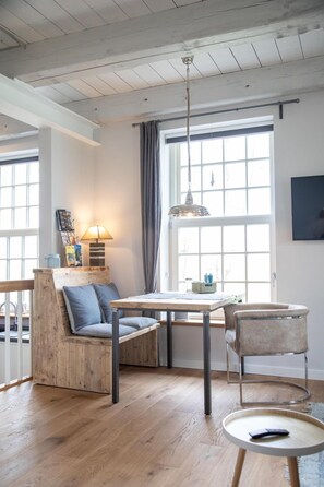 A cozy room with a wooden bench, a table, and chairs. There is a window with curtains, a hanging light, and a picture on the wall.